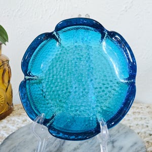 May include: A round, turquoise glass dish with a textured surface and a dark blue scalloped rim. The dish is displayed on a clear stand, showcasing its unique shape and color. The dish is likely vintage or retro.