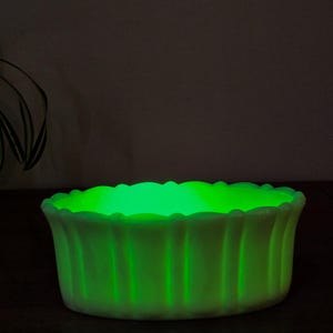 Op de afbeelding: Een gloeiende, lichtgroene glazen kom met een geschulpte rand en verticale geribbelde zijkanten. De kom straalt van binnenuit een heldergroen licht uit, wat een opvallend visueel effect creëert. De kom is ongeveer 15 cm in diameter en 7,6 cm hoog.