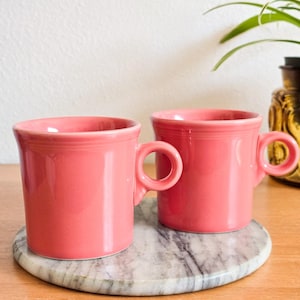 Puede incluir: Dos tazas de cerámica de color coral con asas redondeadas. Las tazas están colocadas sobre un posavasos redondo con diseño de mármol. Las tazas tienen un diseño sencillo y clásico y probablemente son para bebidas.