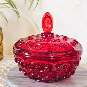 May include: A vintage, ruby red glass candy dish with a lid. The dish has a textured, oval pattern and a matching oval handle on the lid. The candy dish is sitting on a marble surface. The overall theme is vintage home decor.