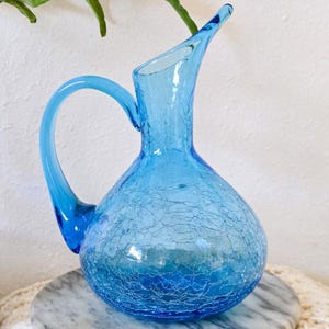 Puede incluir: Una jarra de vidrio azul claro vintage con acabado craquelado. La jarra tiene un cuerpo redondeado, un asa curva y un pico. La jarra está sobre una superficie de mármol. Es un artículo decorativo.