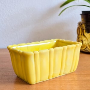 Puede incluir: Una jardinera rectangular de cerámica amarilla brillante con un borde festoneado y lados acanalados verticales. La jardinera está vacía y se encuentra sobre una superficie de madera. Una botella decorativa de vidrio ámbar está en el fondo.