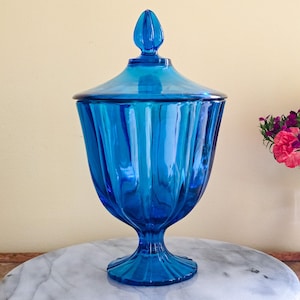 May include: A vibrant blue glass jar with a lid, featuring a fluted design. The jar has a pedestal base and a decorative finial on the lid. The jar is displayed on a marble surface.