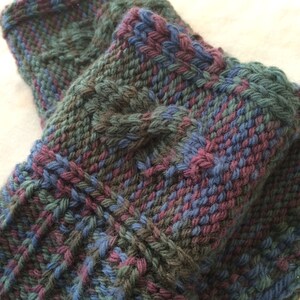 May include: Close-up of a knitted fabric with a multi-colored pattern in shades of blue, green, and purple. The fabric has a textured, cable-knit design.