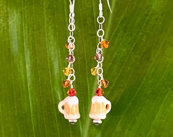 May include: A pair of silver dangle earrings with a ceramic beer mug charm. The earrings feature a chain of small, colorful crystals in shades of orange, red, and brown.