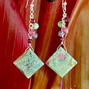 Peut inclure: Une paire de boucles d'oreilles en argent avec des gouttes de pierres précieuses vertes et roses. Les boucles d'oreilles présentent une pierre verte de forme carrée avec des taches roses et blanches, et une chaîne en argent avec de petites perles vertes, roses et bleues.