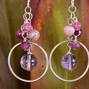 Peut inclure: Boucles d'oreilles créoles en argent avec une pierre précieuse améthyste pendante et un groupe de perles roses et violettes.