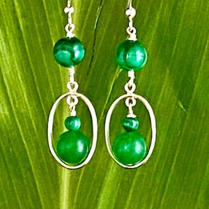 May include: A pair of green malachite gemstone earrings with silver hoops. The earrings feature a round malachite stone at the top and a larger round malachite stone hanging from a silver hoop.
