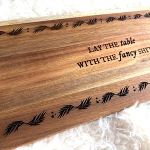 May include: Wooden serving board with the text "LAY THE TABLE WITH THE FANCY SHIT" and decorative leaf and star designs. The board is made of light brown wood.