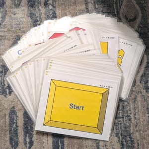 May include: A stack of white cards with yellow and red geometric designs and the word "Start" in blue text. The cards are in clear plastic sleeves. Some cards have the text "LA, X, M" and numbers.