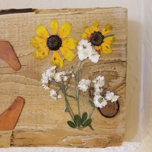 May include: A rustic wooden sign with pressed yellow and white flowers. The flowers are arranged in a bouquet and are set in resin.