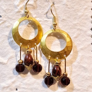 May include: Gold-toned hoop earrings with a hammered texture. Each earring features a dangling design with a dark red, oval-shaped bead, a smaller gold bead, and a faceted, dark red bead. The earrings have a hook closure.