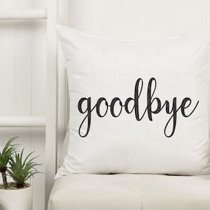 May include: A white pillow with the word "goodbye" written in black cursive script. The pillow is sitting on a white couch with a white blanket draped over the back.