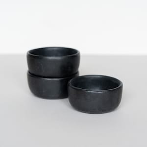 Puede incluir: Tres cuencos de cerámica negra apilados uno encima del otro. Los cuencos son redondos y tienen un acabado mate.