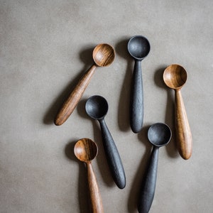 May include: Six wooden spoons, three are natural wood and three are black stained. The spoons are arranged in a fan shape on a grey background.