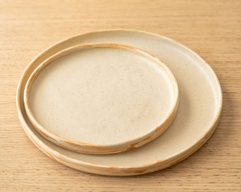 May include: Two beige ceramic plates with a speckled finish and a light brown rim. The plates are nested together on a wooden surface.