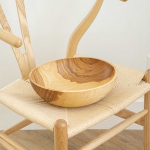 May include: A wooden bowl with a natural wood grain pattern sits on a woven chair seat. The chair is made of light wood and has a curved back.