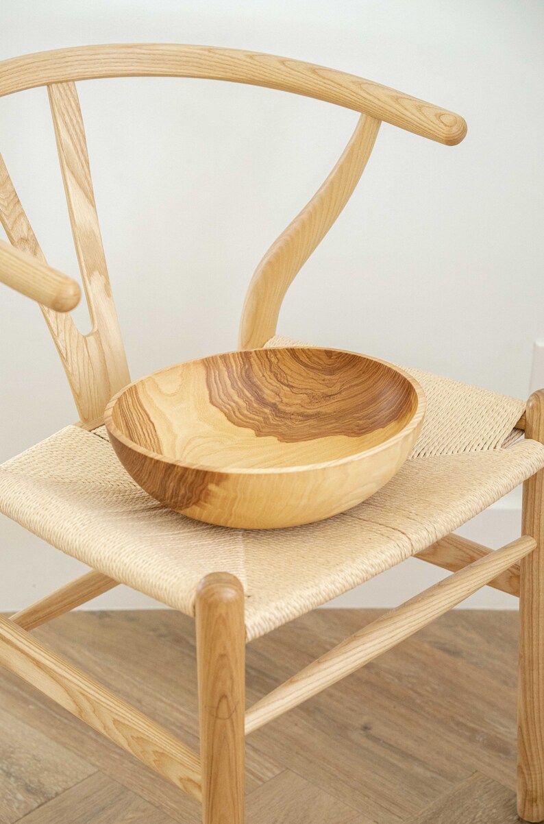 May include: A wooden bowl with a natural wood grain pattern sits on a woven chair seat. The chair is made of light wood and has a curved back.