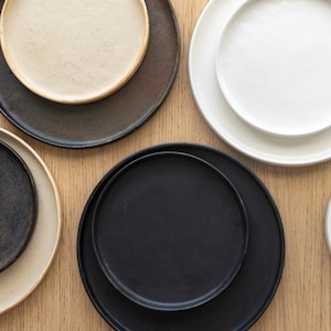 Puede incluir: Vista cenital de una colecci&oacute;n de platos de cer&aacute;mica de varios tama&ntilde;os y colores. Los platos incluyen tonos negros, blancos y beige. Los platos est&aacute;n dispuestos sobre una superficie de madera clara, mostrando una est&eacute;tica minimalista.