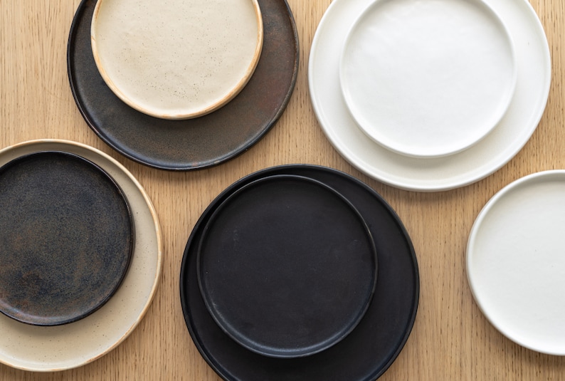Puede incluir: Vista cenital de una colecci&oacute;n de platos de cer&aacute;mica de varios tama&ntilde;os y colores. Los platos incluyen tonos negros, blancos y beige. Los platos est&aacute;n dispuestos sobre una superficie de madera clara, mostrando una est&eacute;tica minimalista.