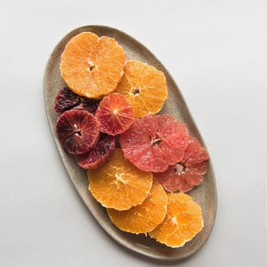 Peut inclure: Un plat ovale en céramique avec un glaçage brun clair contient une variété de tranches d'agrumes. Le plat est rempli de tranches d'orange, de pamplemousse et d'orange sanguine, disposées en dégradé du jaune au rouge.