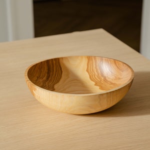 May include: A large, wooden bowl with a natural wood finish. The bowl is made of light-colored wood with dark brown grain patterns. The bowl is sitting on a light-colored wooden table.