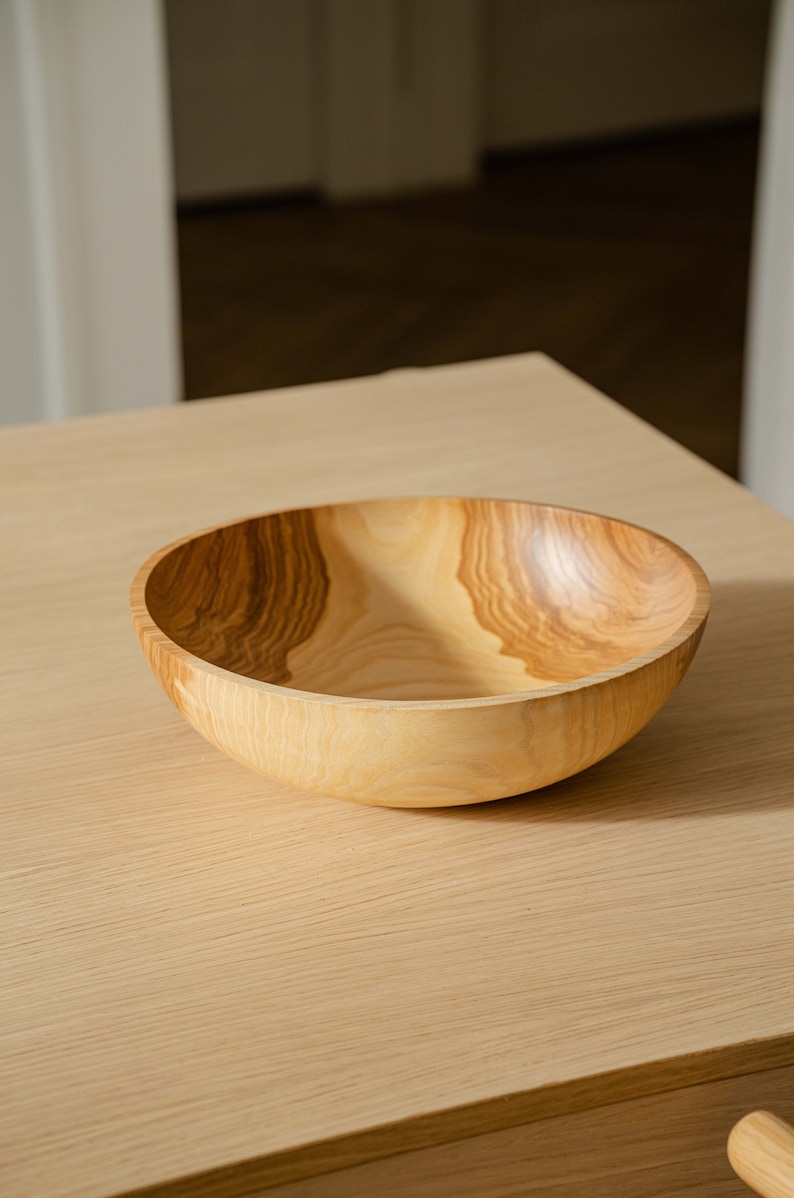 May include: A large, wooden bowl with a natural wood finish. The bowl is made of light-colored wood with dark brown grain patterns. The bowl is sitting on a light-colored wooden table.