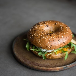 Puede incluir: Un s&aacute;ndwich de bagel con semillas de s&eacute;samo, relleno de r&uacute;cula, un relleno amarillo y una crema blanca, presentado en un plato marr&oacute;n. El bagel es de color marr&oacute;n dorado y el plato tiene un tono terroso. El fondo es gris apagado.