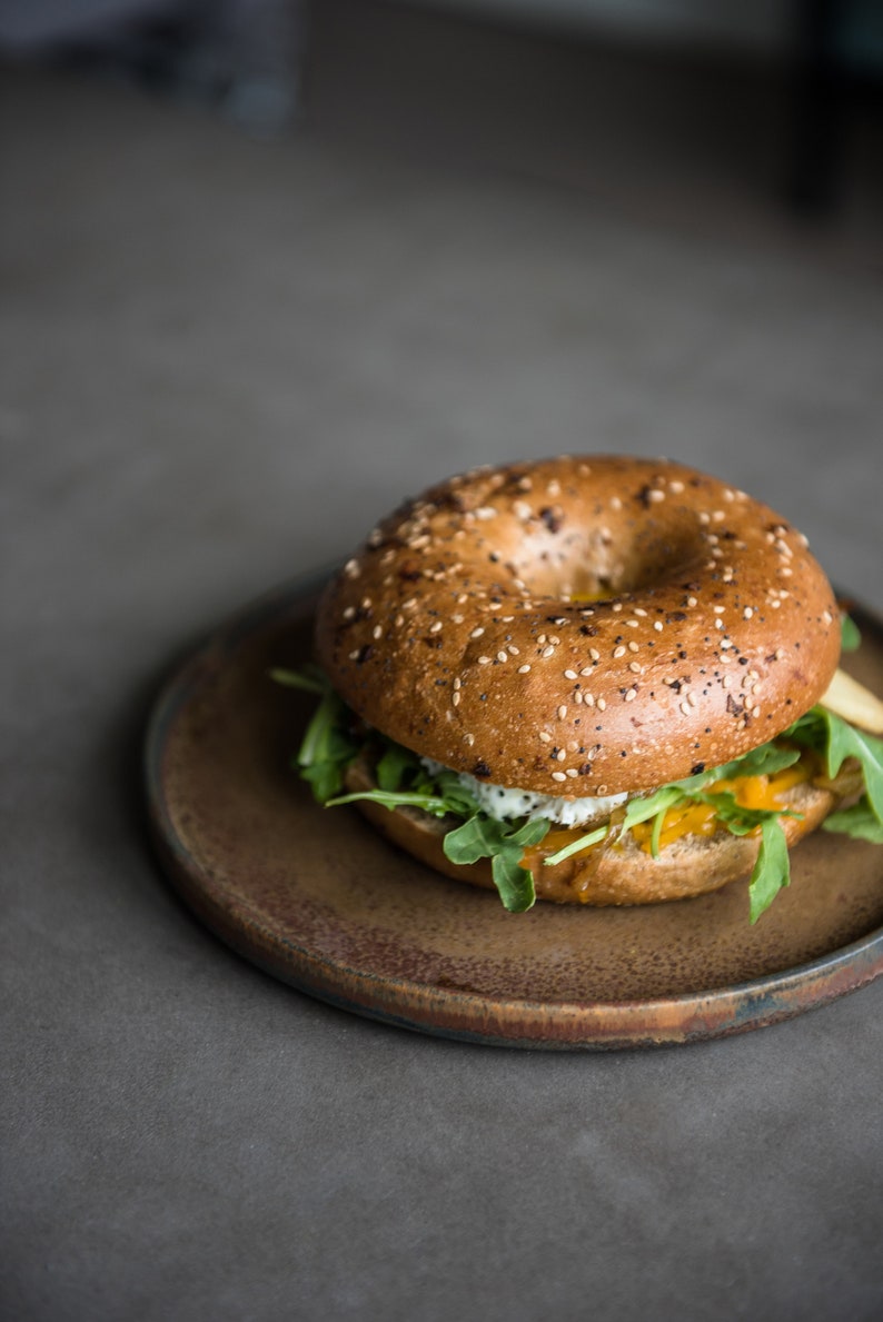 Puede incluir: Un s&aacute;ndwich de bagel con semillas de s&eacute;samo, relleno de r&uacute;cula, un relleno amarillo y una crema blanca, presentado en un plato marr&oacute;n. El bagel es de color marr&oacute;n dorado y el plato tiene un tono terroso. El fondo es gris apagado.