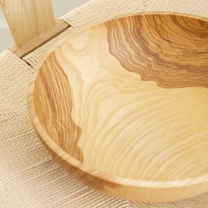 May include: A large, light brown wooden bowl with a natural wood grain pattern. The bowl is sitting on a woven chair seat.