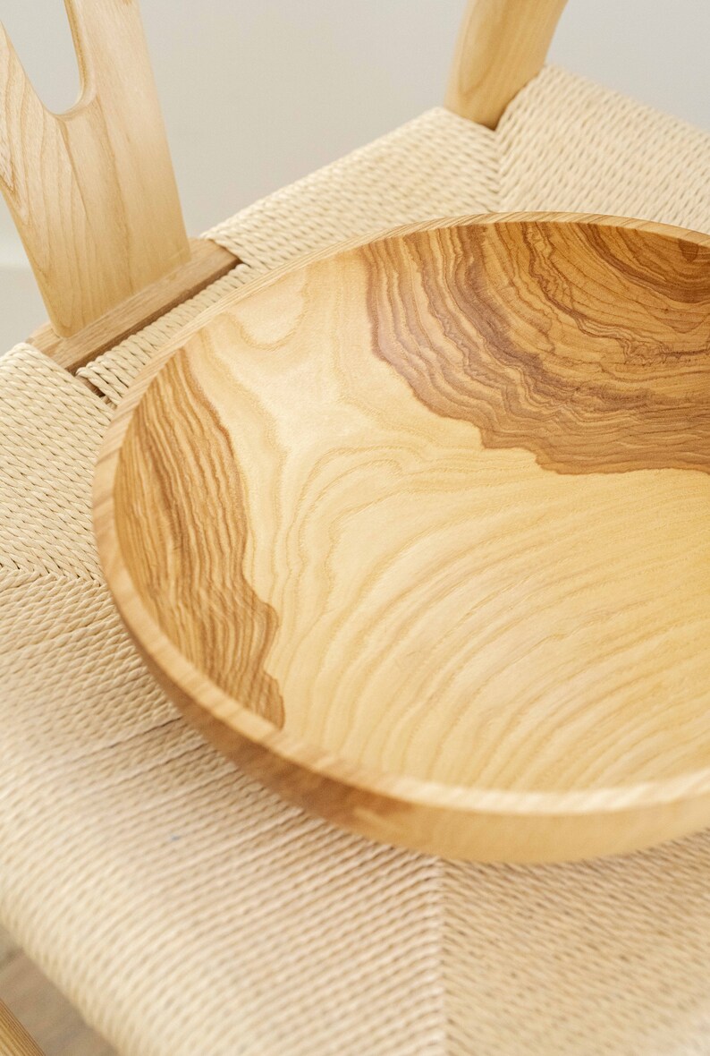 May include: A large, light brown wooden bowl with a natural wood grain pattern. The bowl is sitting on a woven chair seat.