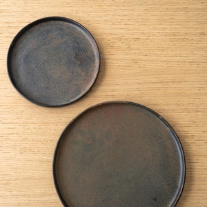 Puede incluir: Dos platos de cer&aacute;mica gris oscuro con un acabado r&uacute;stico y moteado. Los platos son redondos y tienen un borde ligeramente elevado. Los platos se muestran sobre una superficie de madera clara.