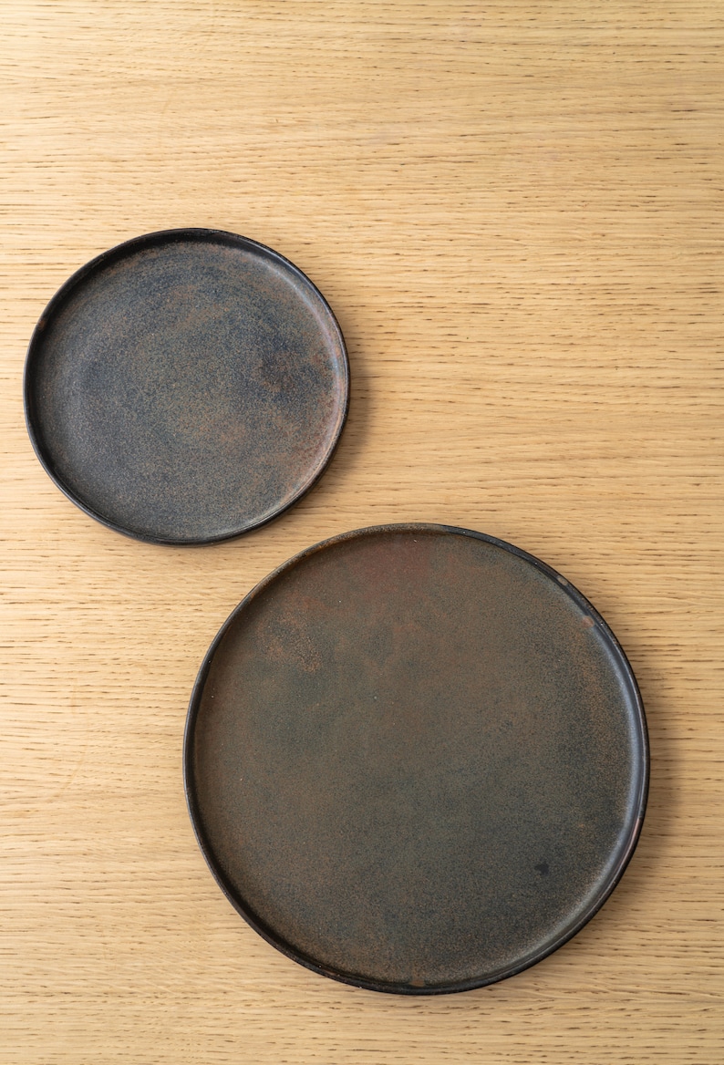 Puede incluir: Dos platos de cer&aacute;mica gris oscuro con un acabado r&uacute;stico y moteado. Los platos son redondos y tienen un borde ligeramente elevado. Los platos se muestran sobre una superficie de madera clara.