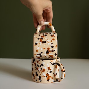 May include: Two ceramic mugs with a beige base and a speckled pattern in brown, black, and white. One mug has a handle on top, and the other has a curved handle on the side. The mugs are stacked on top of each other.