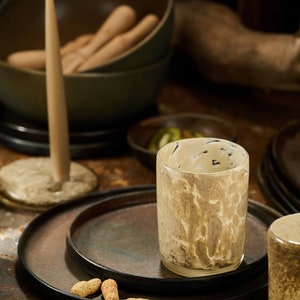 Puede incluir: Una vela encendida, un vaso decorativo con un dise&ntilde;o veteado y varios platos oscuros est&aacute;n colocados sobre una mesa. Tambi&eacute;n se ven cuencos y cacahuetes, creando un ambiente c&aacute;lido.