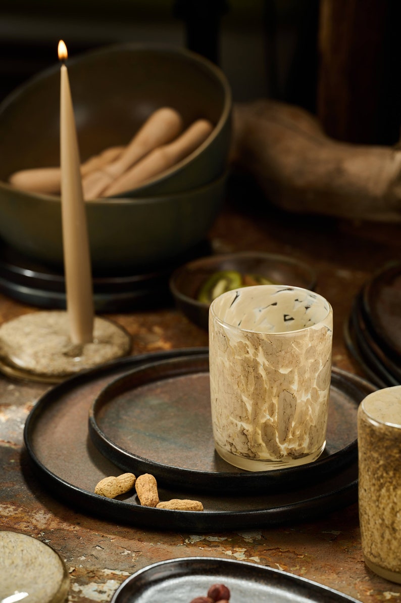 Puede incluir: Una vela encendida, un vaso decorativo con un dise&ntilde;o veteado y varios platos oscuros est&aacute;n colocados sobre una mesa. Tambi&eacute;n se ven cuencos y cacahuetes, creando un ambiente c&aacute;lido.
