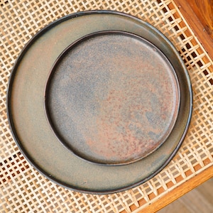 Puede incluir: Dos platos de cer&aacute;mica, uno dentro del otro, con un acabado r&uacute;stico y moteado en tonos marr&oacute;n, verde y bronce. Los platos est&aacute;n colocados sobre una silla de mimbre trenzado, creando una est&eacute;tica natural. Ideales para servir alimentos.