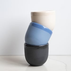 May include: A stack of three ceramic cups with rounded bases. The top cup is cream, the middle cup is a light blue with a glossy rim, and the bottom cup is a matte charcoal gray with a glossy rim. Each cup features a smooth, simple design.