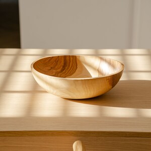 May include: A light brown wooden bowl sits on a light wood table with sunlight streaming through a window. The bowl is empty and has a smooth, rounded shape.