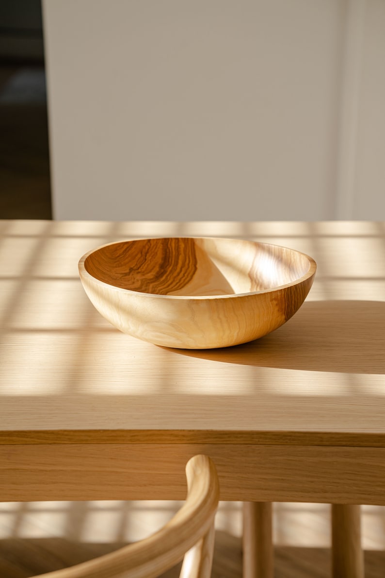 May include: A light brown wooden bowl sits on a light wood table with sunlight streaming through a window. The bowl is empty and has a smooth, rounded shape.