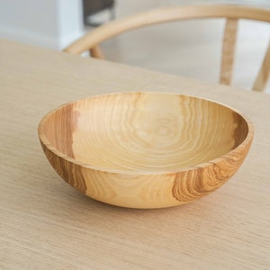 May include: A large, light brown wooden bowl with a smooth, rounded interior. The bowl is made of natural wood with visible grain patterns.