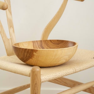 May include: A large, wooden bowl sits on a woven chair seat. The bowl is made of light brown wood with darker brown grain patterns. The chair is made of light brown wood with a woven seat.