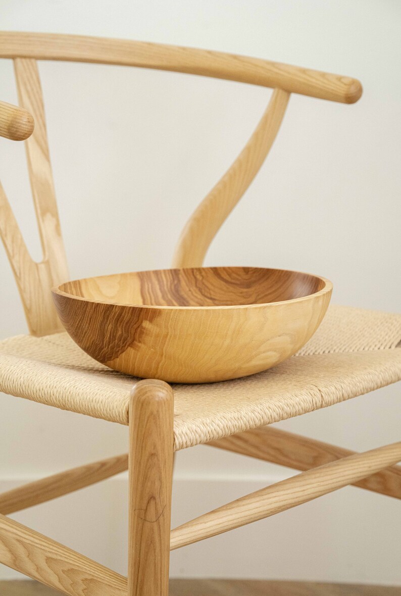 May include: A large, wooden bowl sits on a woven chair seat. The bowl is made of light brown wood with darker brown grain patterns. The chair is made of light brown wood with a woven seat.