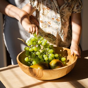 May include: A child reaches for a bunch of green grapes in a wooden bowl with two pears. The bowl is on a light wood table.