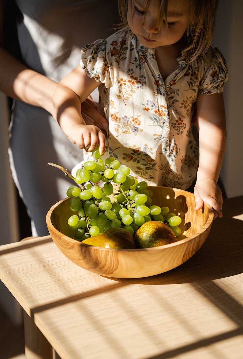 May include: A child reaches for a bunch of green grapes in a wooden bowl with two pears. The bowl is on a light wood table.