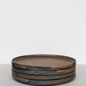 Puede incluir: Una pila de cuatro platos de cer&aacute;mica r&uacute;sticos. Los platos tienen un dise&ntilde;o plano y redondo con un centro marr&oacute;n oscuro y un borde azul-verdoso y marr&oacute;n. Los platos est&aacute;n apilados uno encima del otro, creando una presentaci&oacute;n simple y elegante.