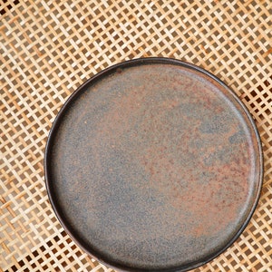 Puede incluir: Un plato redondo de estilo r&uacute;stico con una superficie oscura y moteada en tonos grises y marrones. El plato tiene un borde ligeramente elevado y se asienta sobre una superficie tejida de color marr&oacute;n claro. Ideal para servir alimentos.