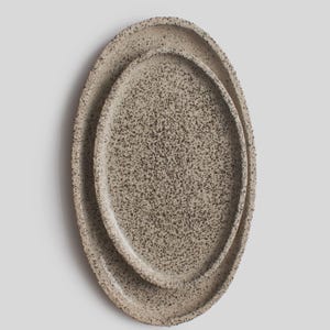 Puede incluir: Dos fuentes ovaladas de cerámica, una dentro de la otra. Ambas tienen un patrón moteado de puntos marrón oscuro sobre un fondo beige claro. Los bordes son elevados y texturizados. Adecuadas para servir alimentos o como elementos decorativos.