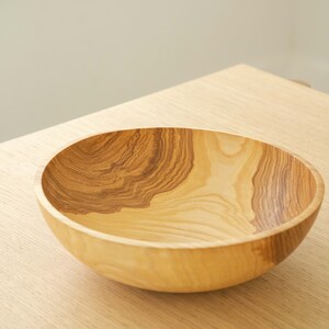 May include: A large, wooden bowl with a natural wood grain pattern. The bowl is empty and sits on a light wood surface.