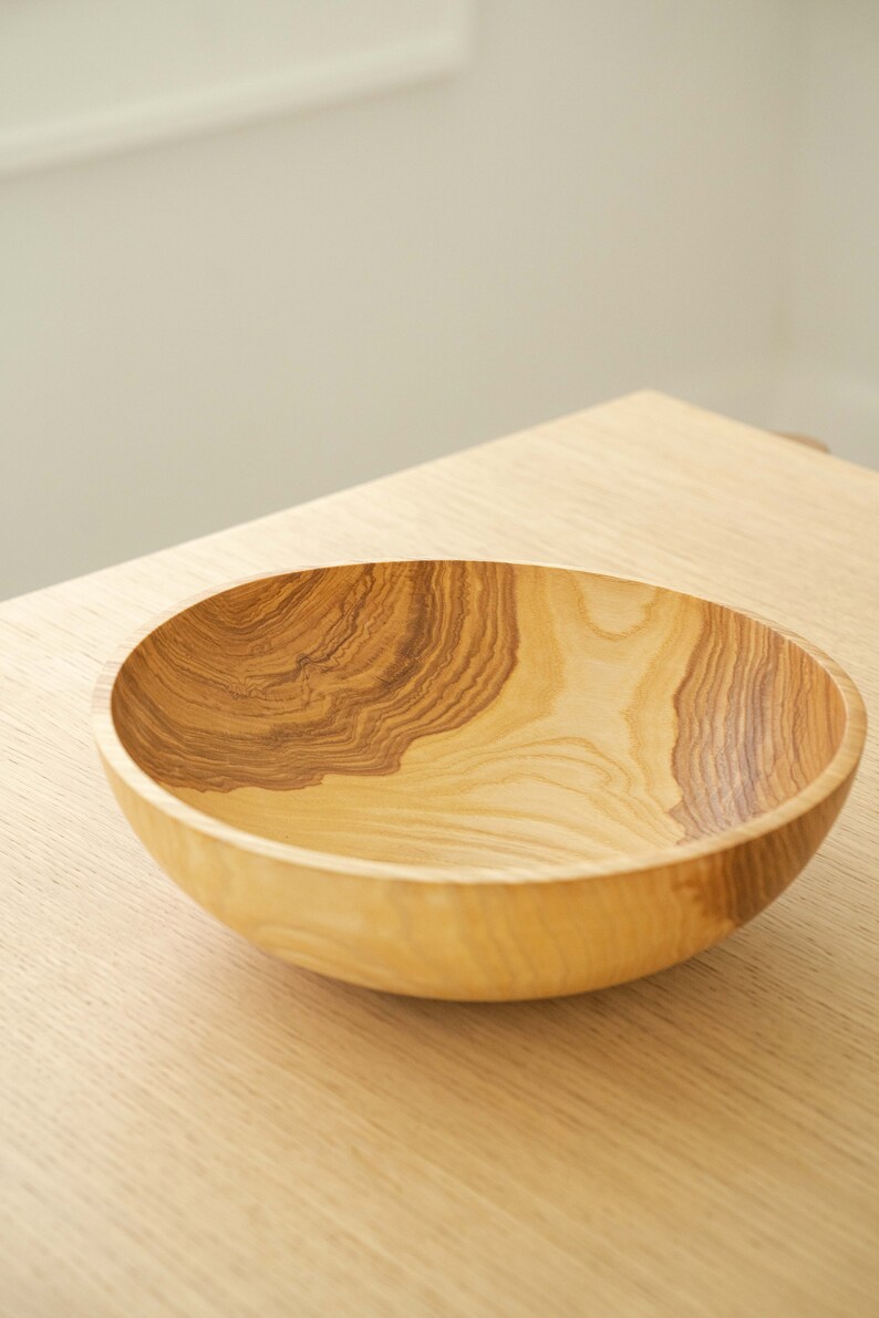 May include: A large, wooden bowl with a natural wood grain pattern. The bowl is empty and sits on a light wood surface.